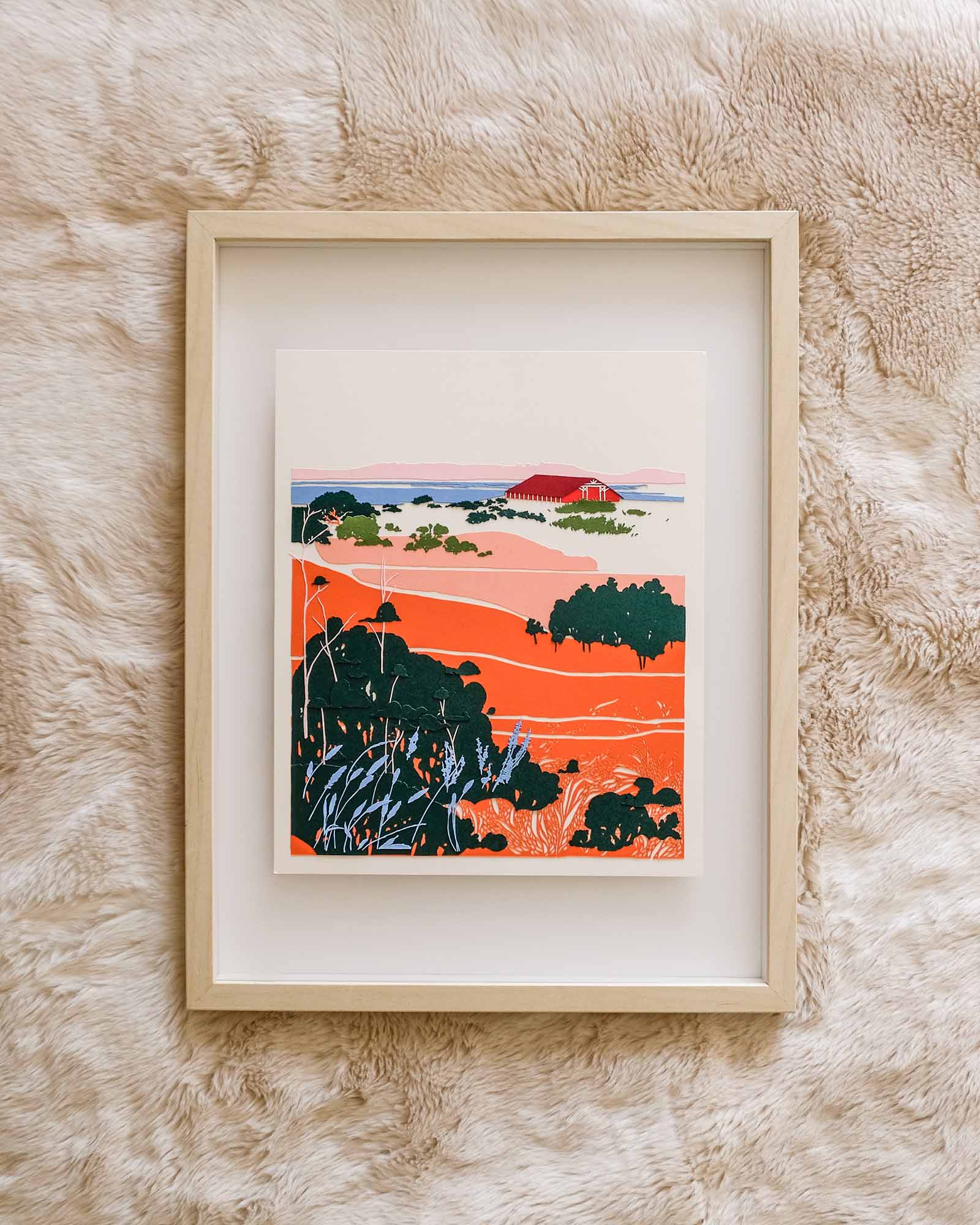 Floating in a light wood frame, sections of organic orange and pink paper shapes are carved with grass-like textures. In the distance, a red paper barn sits on a shore by a blue lake, where hazy pink hills are visible on the horizon. The scene is punctuated by layers of green brush and strands of blue grasses. The entire piece sits against a white fur, which accentuates the movements of the grasses in the artwork.