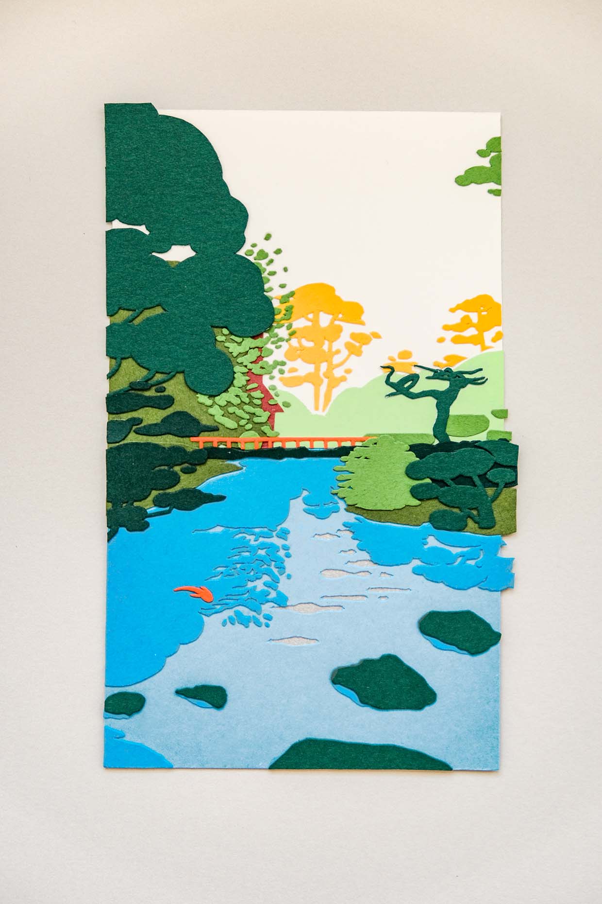 A bright blue paper pond reflects foliage of jade and forest and grass green against a white sky. Hints of orange appear as a fish and manmade structures peeking out from a distance.