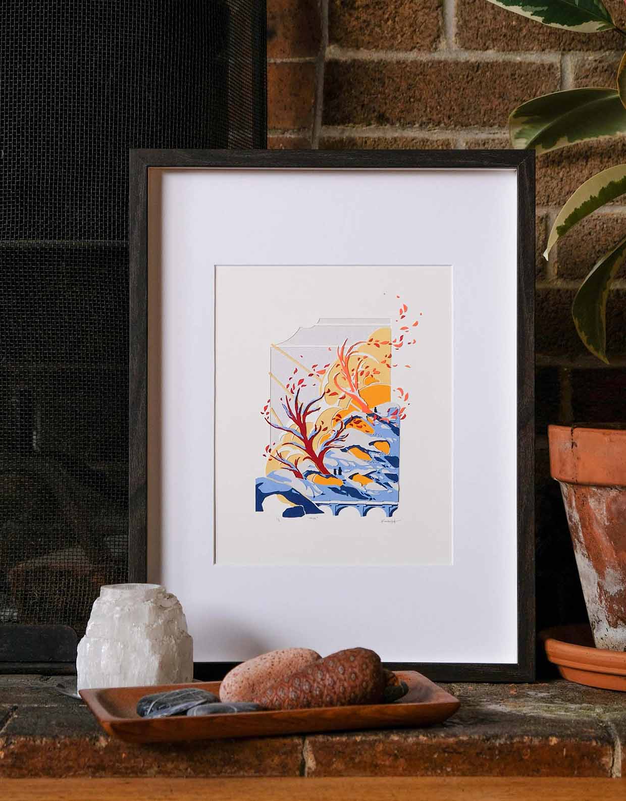 The artwork is matted, framed, and displayed against a brick fireplace with various objects like rocks, pinecones, and candles.