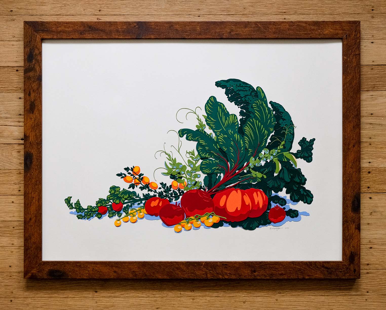 A bounty of paper produce tumbles off the page, including kale, chard, sweet peas, various tomatoes, beets, and kumquats. The greens, reds, and oranges are offset by a blue paper cast shadow. The full artwork is surrounded by a dark wood frame against a lighter but worn wood background.