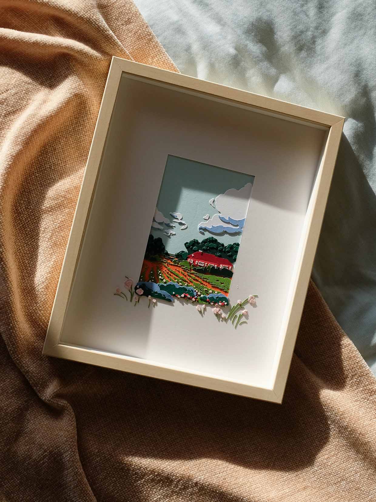 The artwork is framed in a light wood frame against light blue and sand-colored fabrics. Strong sunlight gives the artwork a feeling of late spring.