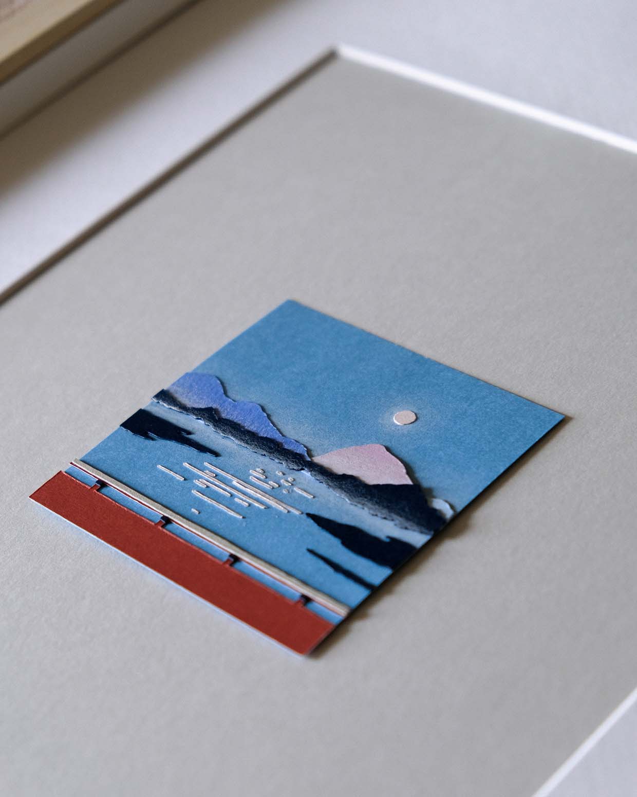 Various shades of cool papers, with vague shapes of hills and land, float above a square of blue paper. A small moon floats above and thin lines like the moon's reflection dance below. The artwork sits on a gray paper background surrounded by a white frame.