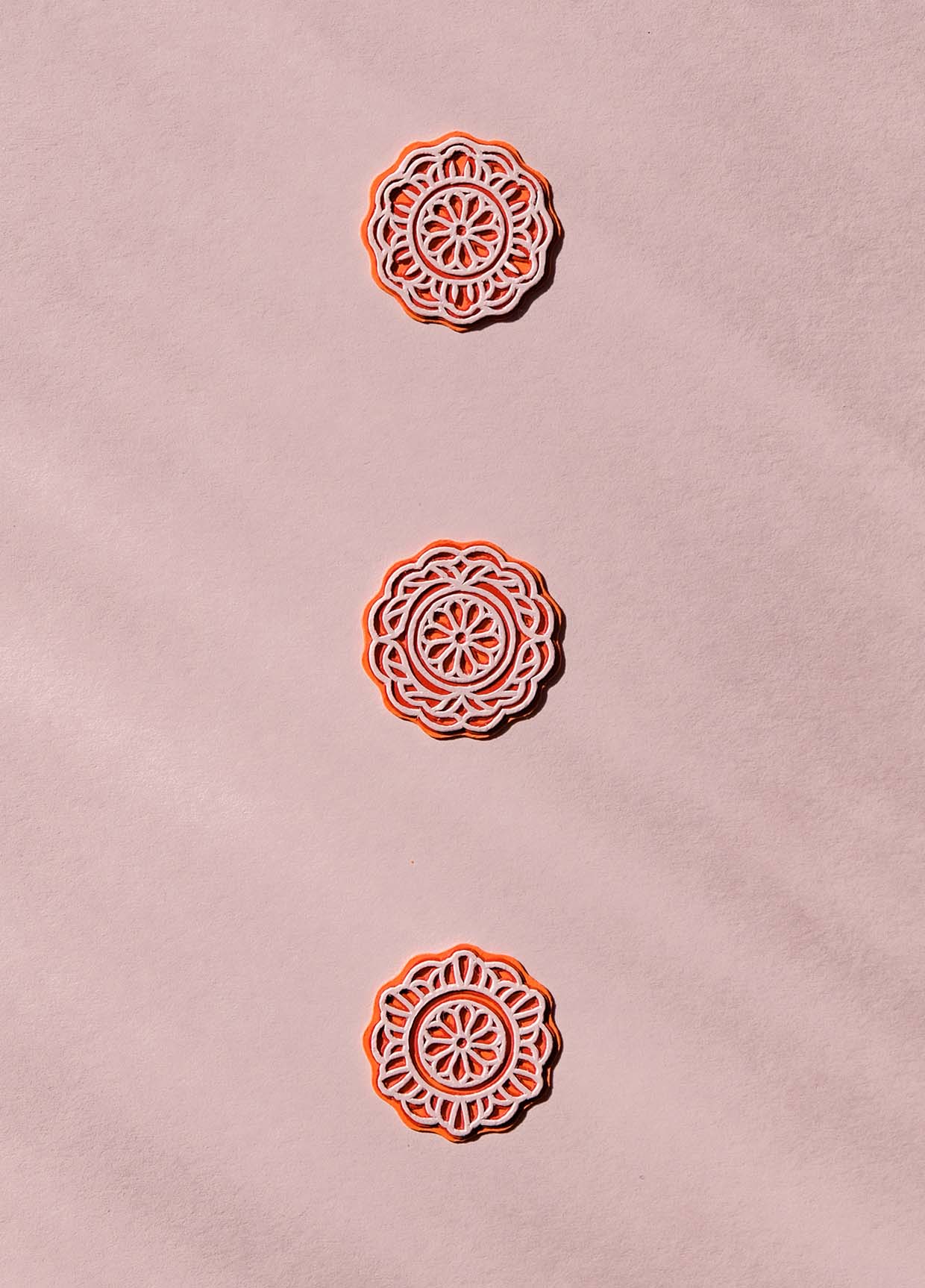 Three orange circles with intricate pink ornamentation vaguely represent the patterns on top of mooncakes. The three pieces float above a light pink paper.