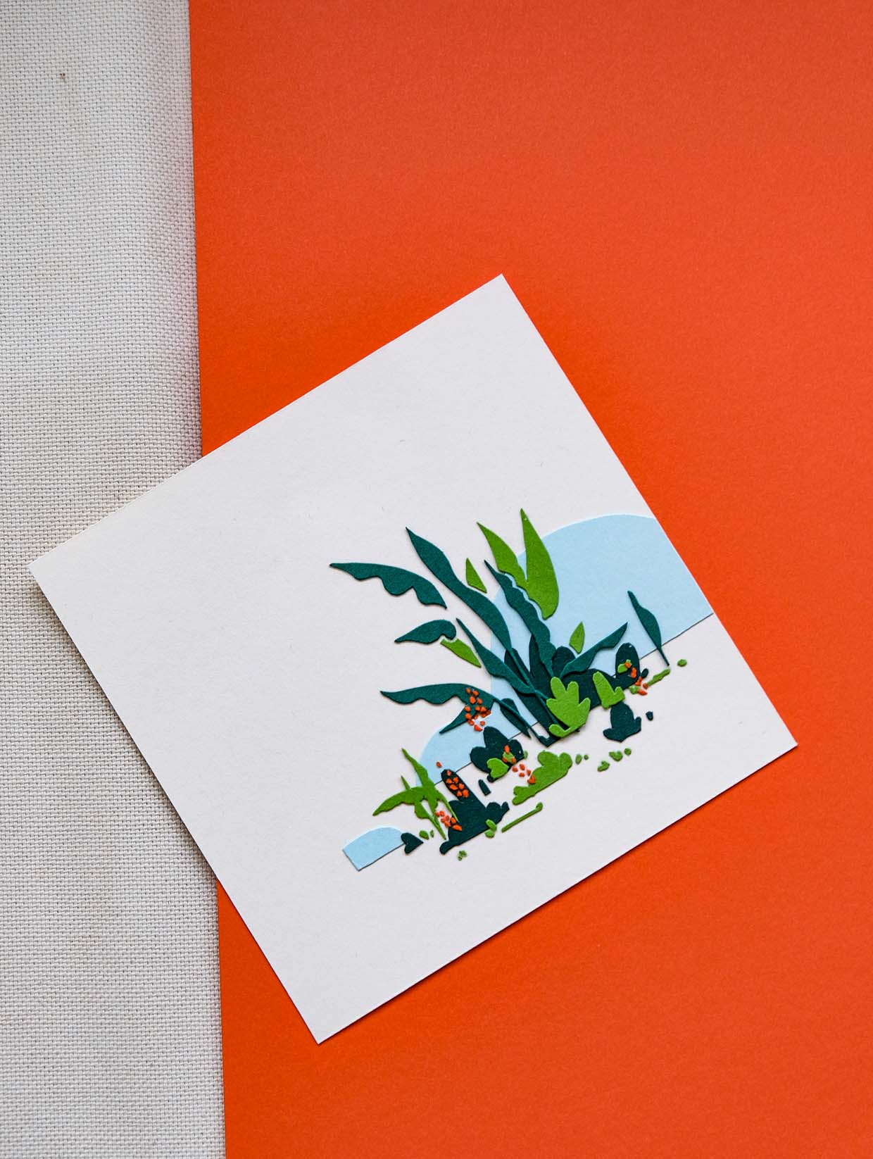 Green abstract leaves and plants are punctuated by tiny orange flower-like elements. Behind them are two blue abstracted hill on a white expanse. The square artwork lies on an angle above a sheet of orange paper and a swatch of white fabric.