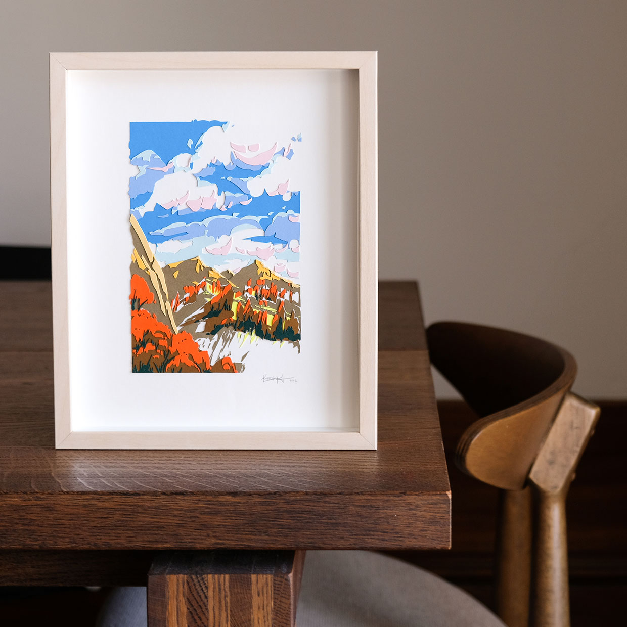 Colorful paper trees, mountains, and a sky full of clouds, framed in light wood, sit on a dark wood table. The colors are reminiscent of autumn leaves under a crisp, chilly sky. The artwork stands on a wood table.