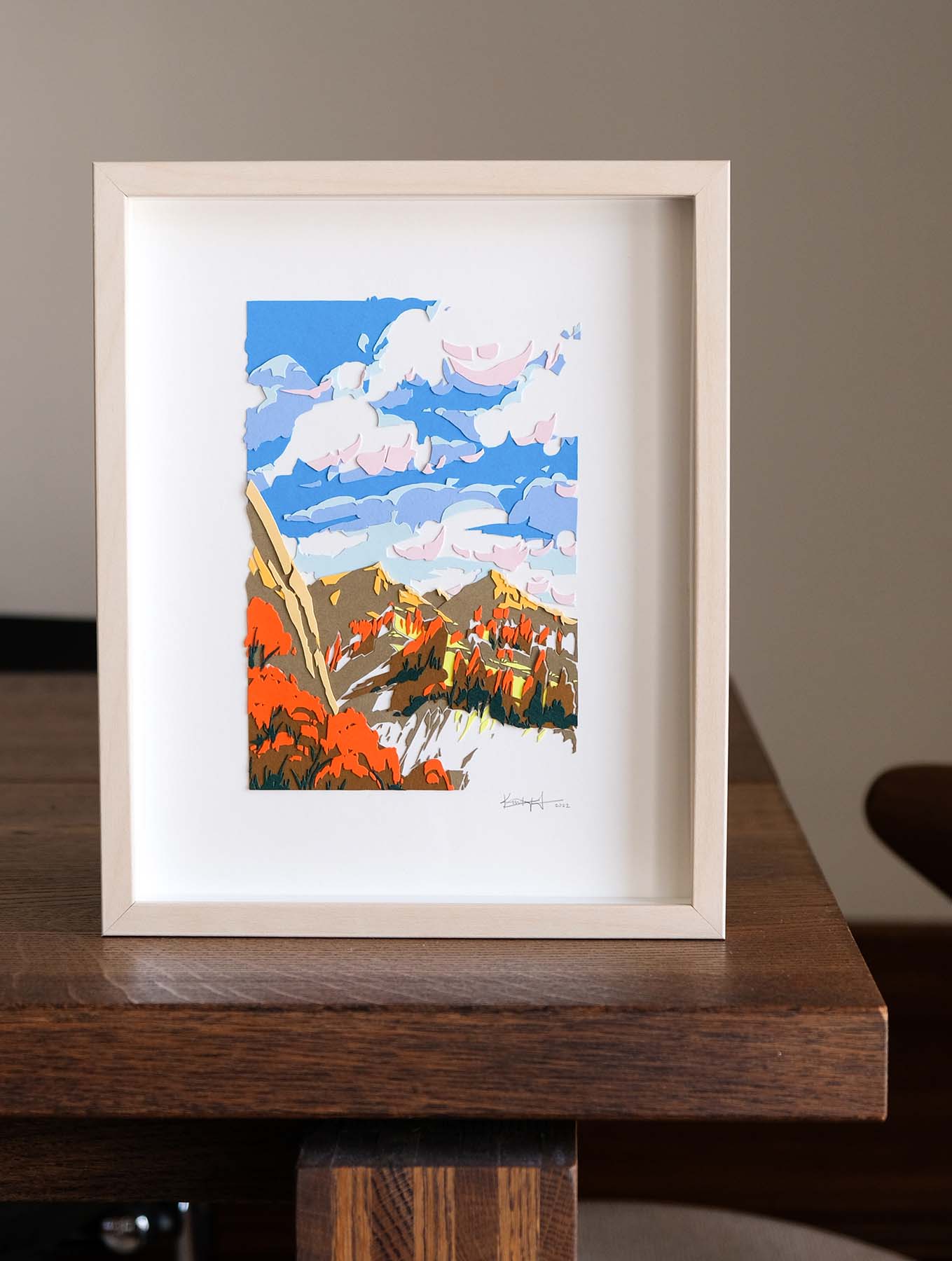 Colorful paper trees, mountains, and a sky full of clouds, framed in light wood, sit on a dark wood table. The colors are reminiscent of autumn leaves under a crisp, chilly sky. The artwork stands on a wood table.