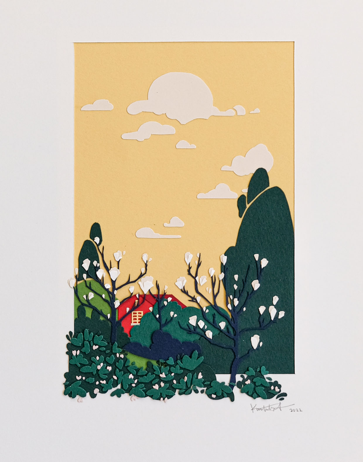 Layers of cut paper depict a red house against a warm yellow sky, behind a grove of magnolia blossoms and other trees. It sits in a white mat, with some of the trees and plants spilling out of the mat opening.