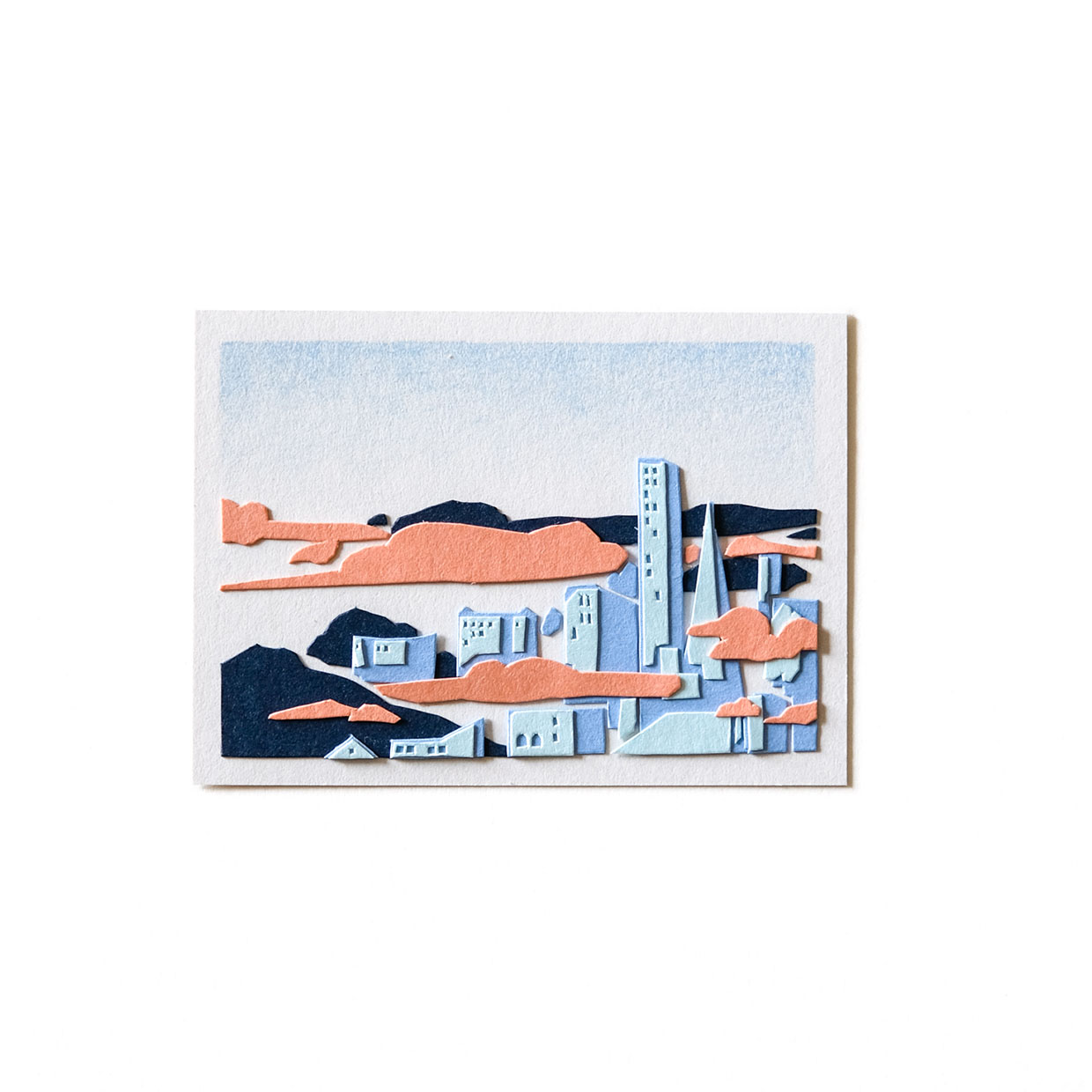 A small piece of cut paper artwork depicts San Francisco’s iconic downtown skyline with surrounded by pink fog and dark blue hills under a light blue sky.