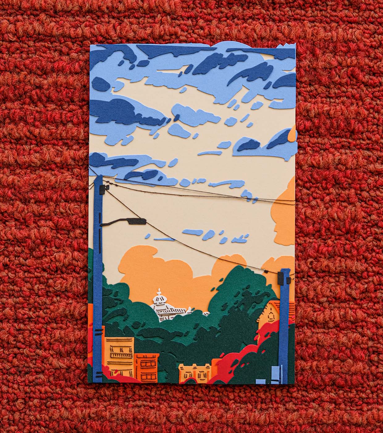 Layers of red, orange, green, and blue paper create a scene of apartment blocks against the green tree tops of Golden Gate Park. Power lines are in the foreground and the Conservatory of Flowers sits in the background against a pale yellow sky with blue clouds. The artwork sits on a rust red rug.