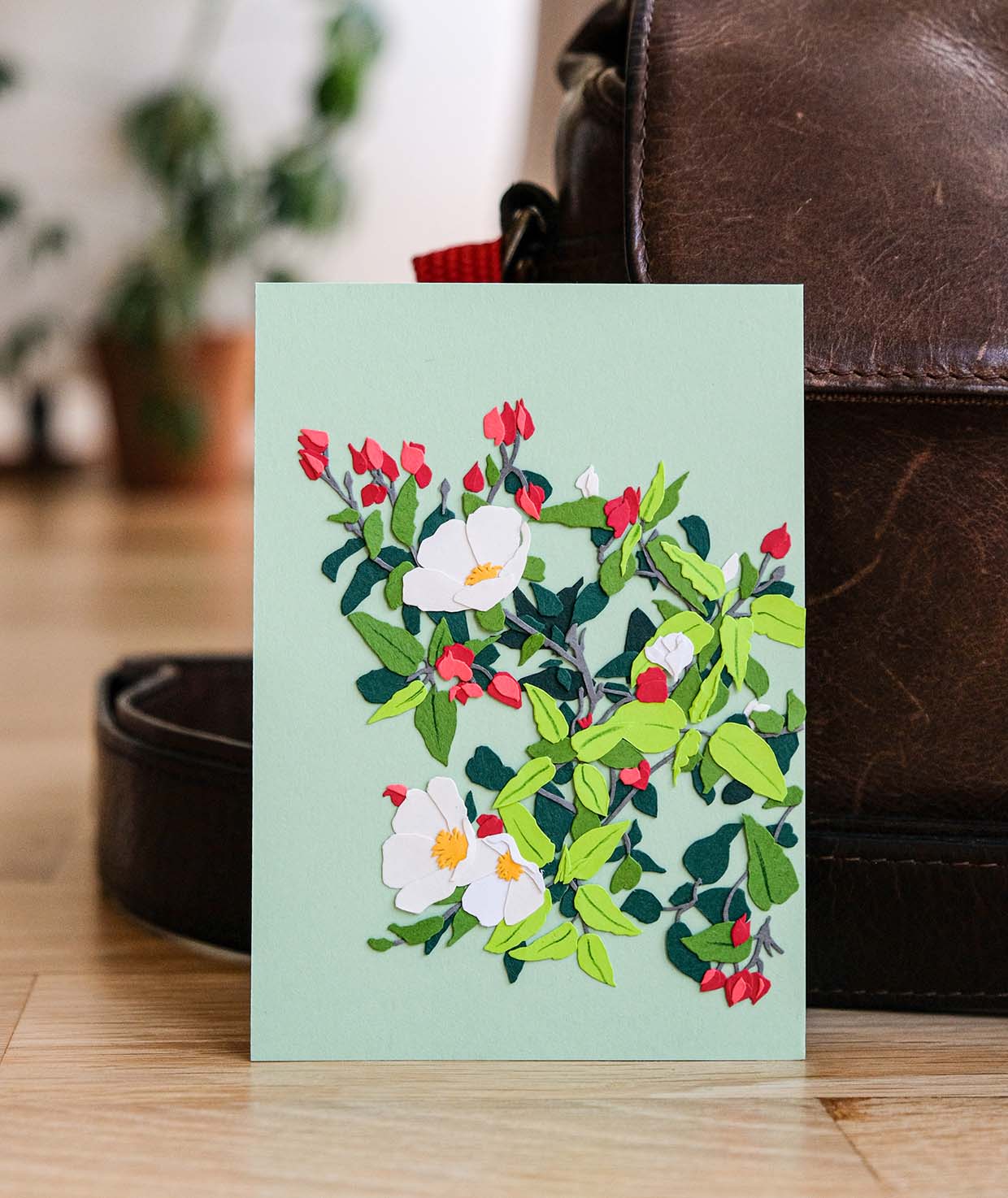 White blooms of salvia cistus are layered on top of red buds, green leaves, and silvery gray stems on a field of light teal. The artwork is made from hand cut paper. Blurred in the background is a wood floor and some real plants.