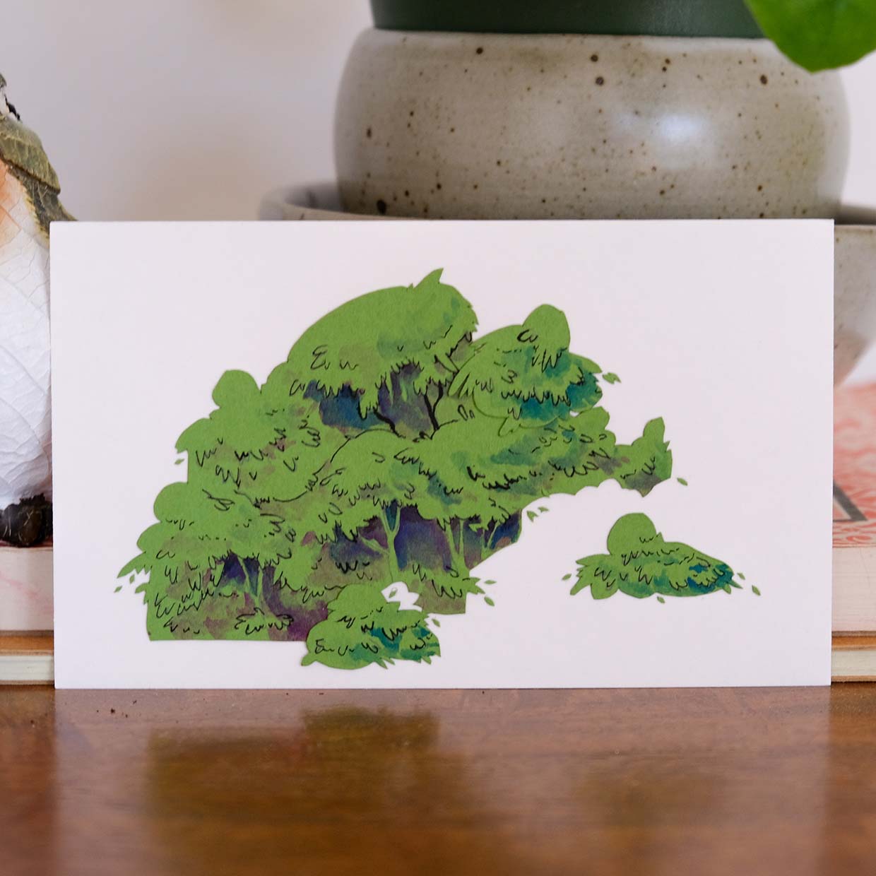 Green paper is cut roughly into the shape of trees with a few leaves scattered around it. Leaf details and canopy shadows are added with colored pencil and ink. The green paper sits on a white background in front of some notebooks and pottery on a desk.