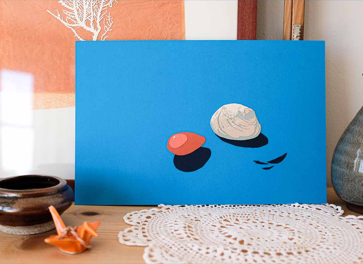 A creamy-colored clam shell and a rusty-colored bulb of kelp, both made of colored paper, sit on a background made of bright teal paper. The artwork is surrounded by white and warm brown curios such as ceramics and a snippet of paper coral.