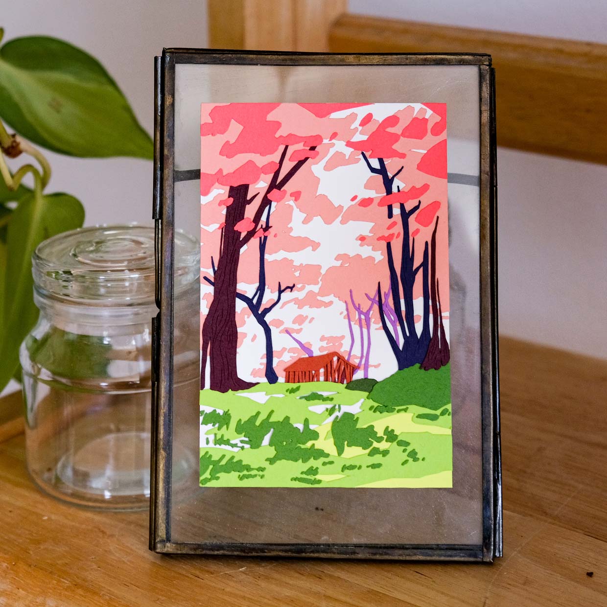 Pink blossoming trees of paper tower over a rundown wooden building in a field of green. The artwork sits in a metal and glass frame next to wood and a vine.