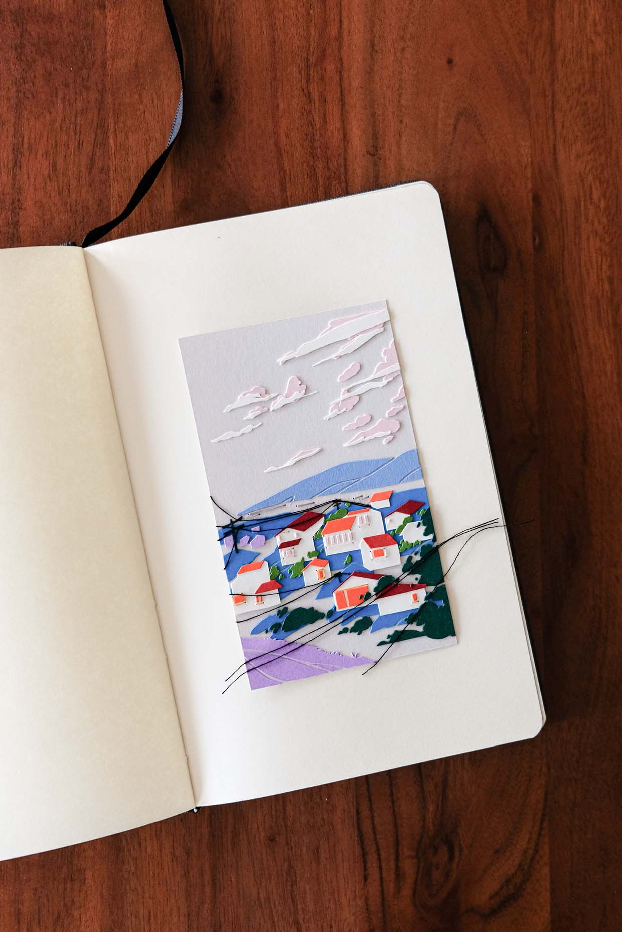 White paper houses with orange and red windows and roofs, sitting among green shrubs and trees, are perched on the slopes of blue hills. White clouds float above them and threads of power lines crisscross before them. The entire artwork rests in a sketchbook lying open on a wood table.