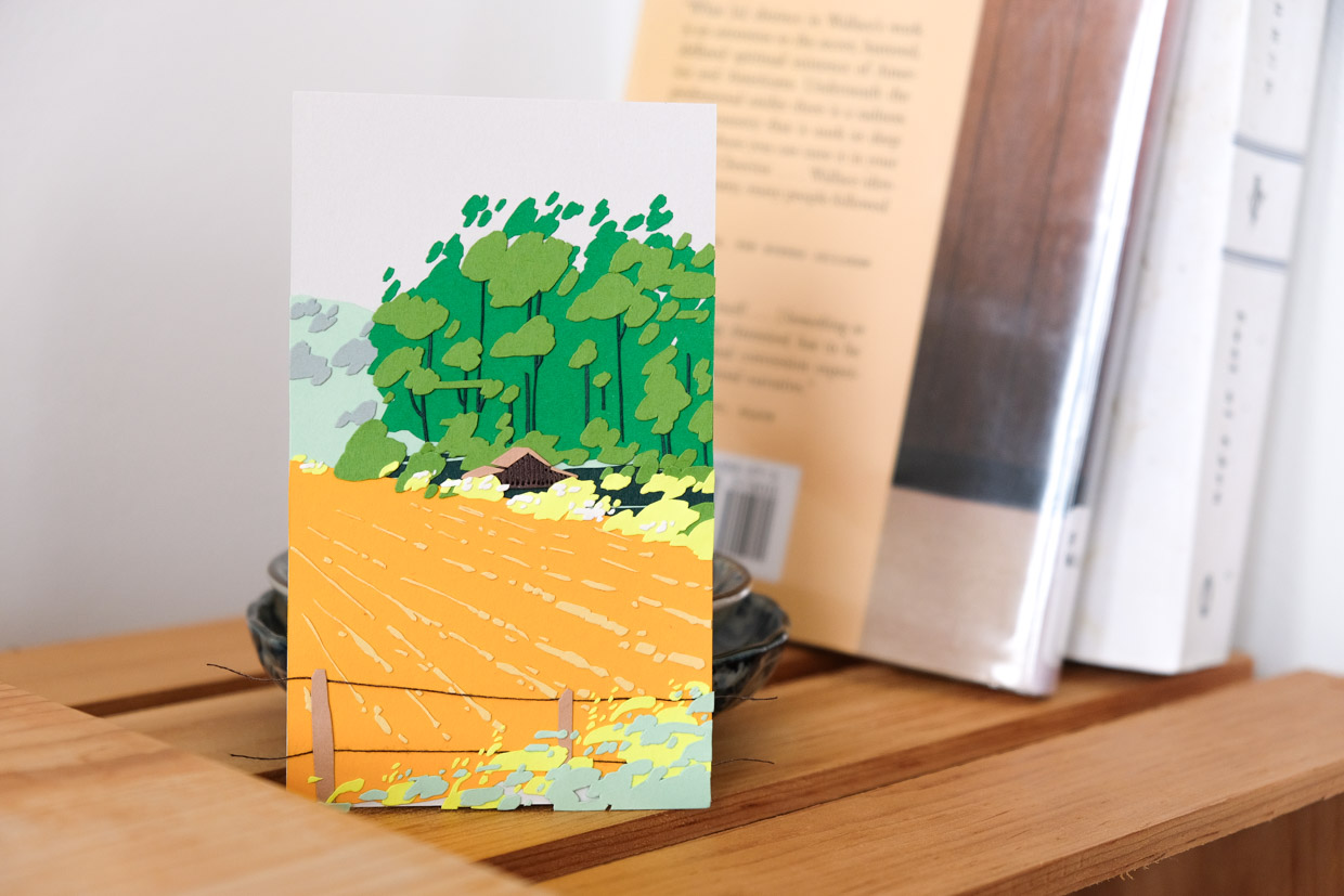 A field of deep goldenrod stretches towards hills on the horizon, skirting past an old barn, shrubs of pale green, and a tall windbreak of deep green eucalyptus. The artwork rests on a wooden shelf next to a couple of books, including Steinbeck’s East of Eden.