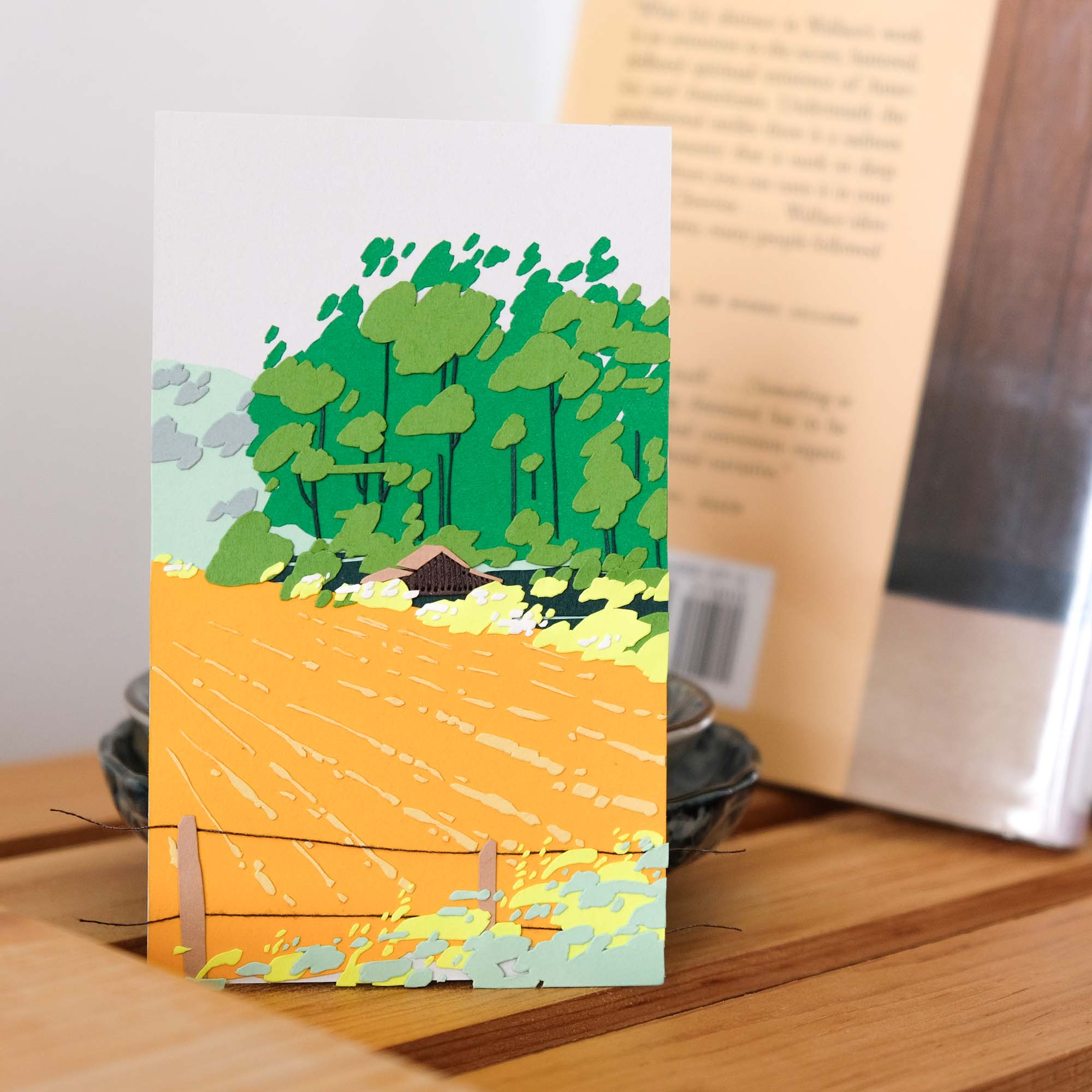 A field of deep goldenrod stretches towards hills on the horizon, skirting past an old barn, shrubs of pale green, and a tall windbreak of deep green eucalyptus. The artwork rests on a wooden shelf next to a couple of books.