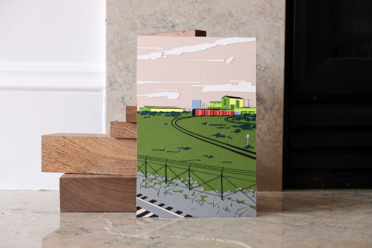 A field of green paper stretches under a dust-colored sky with a green-and-blue hued industrial sprawl on the horizon. A rust-colored train sits on a pair of railroad tracks. The artwork rests next to a stack of wooden blocks on a stone surface.