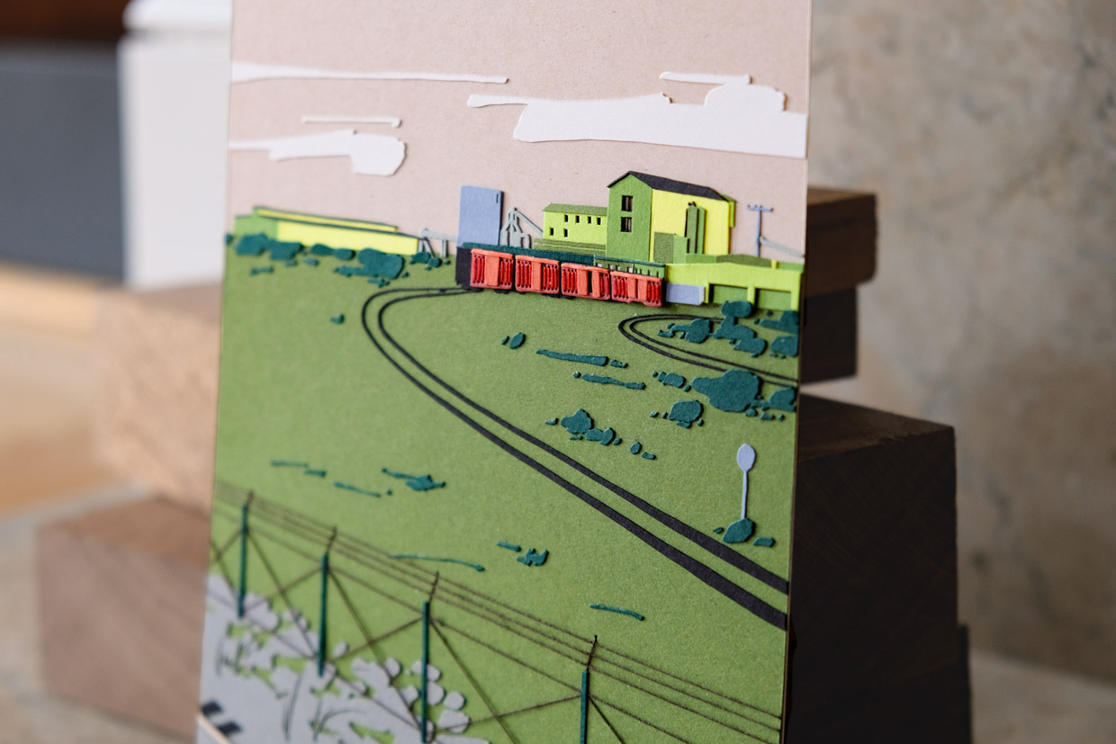 A cropped view of the artwork focuses on the red thread that embellishes the train cars. The layers of green paper around the train are also emphasized by the angle of the light.