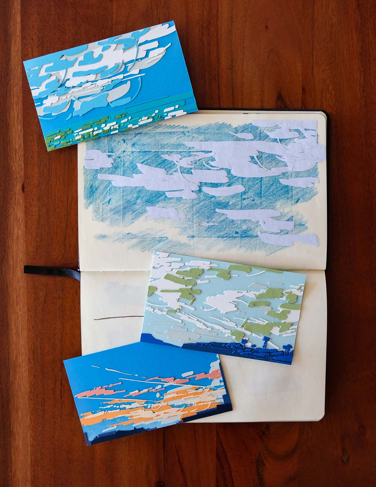 All four pieces from the Clouds series are arranged around a small sketchbook on a wood table.