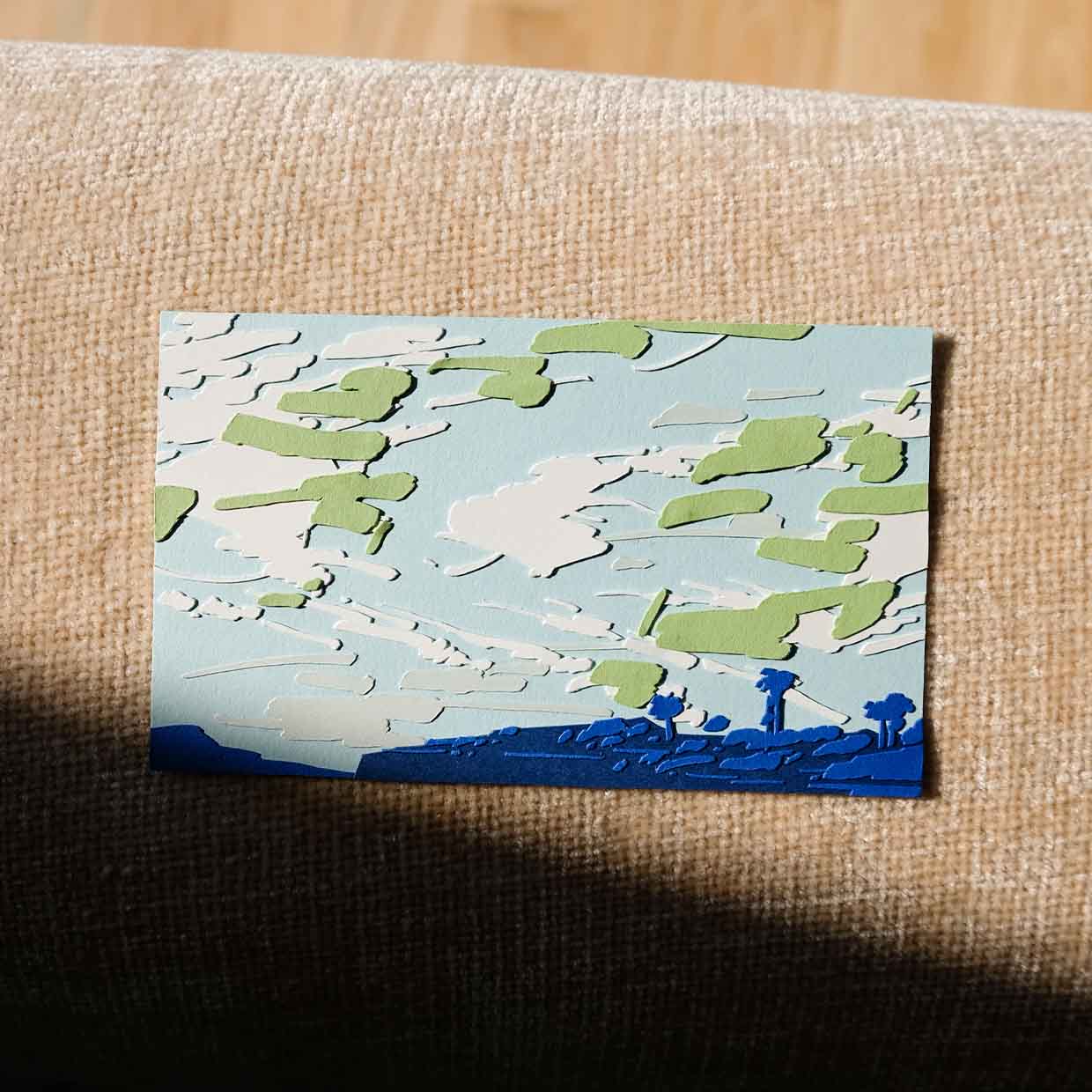 White, gray, and jade green paper clouds are scattered in layers across a light blue sky. Shallow cliffs and trees of indigo line the horizon. The artwork sits on a piece of wood furniture next to a tillandsia.