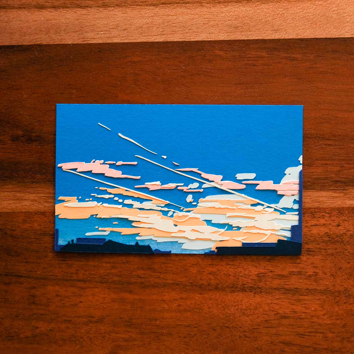 In a rich blue paper sky above indigo rooftops, pale paper clouds of orange, blush, and soft white streak across the sunset sky from bottom right to top left. The artwork floats on a wood surface with undulating lines.