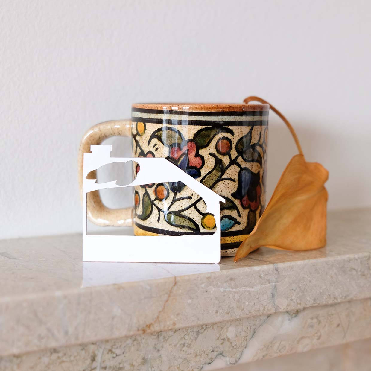 A narrow marble fireplace mantel is adorned by a white paper house, a tiny earthy mug, and a dried philodendron leaf.