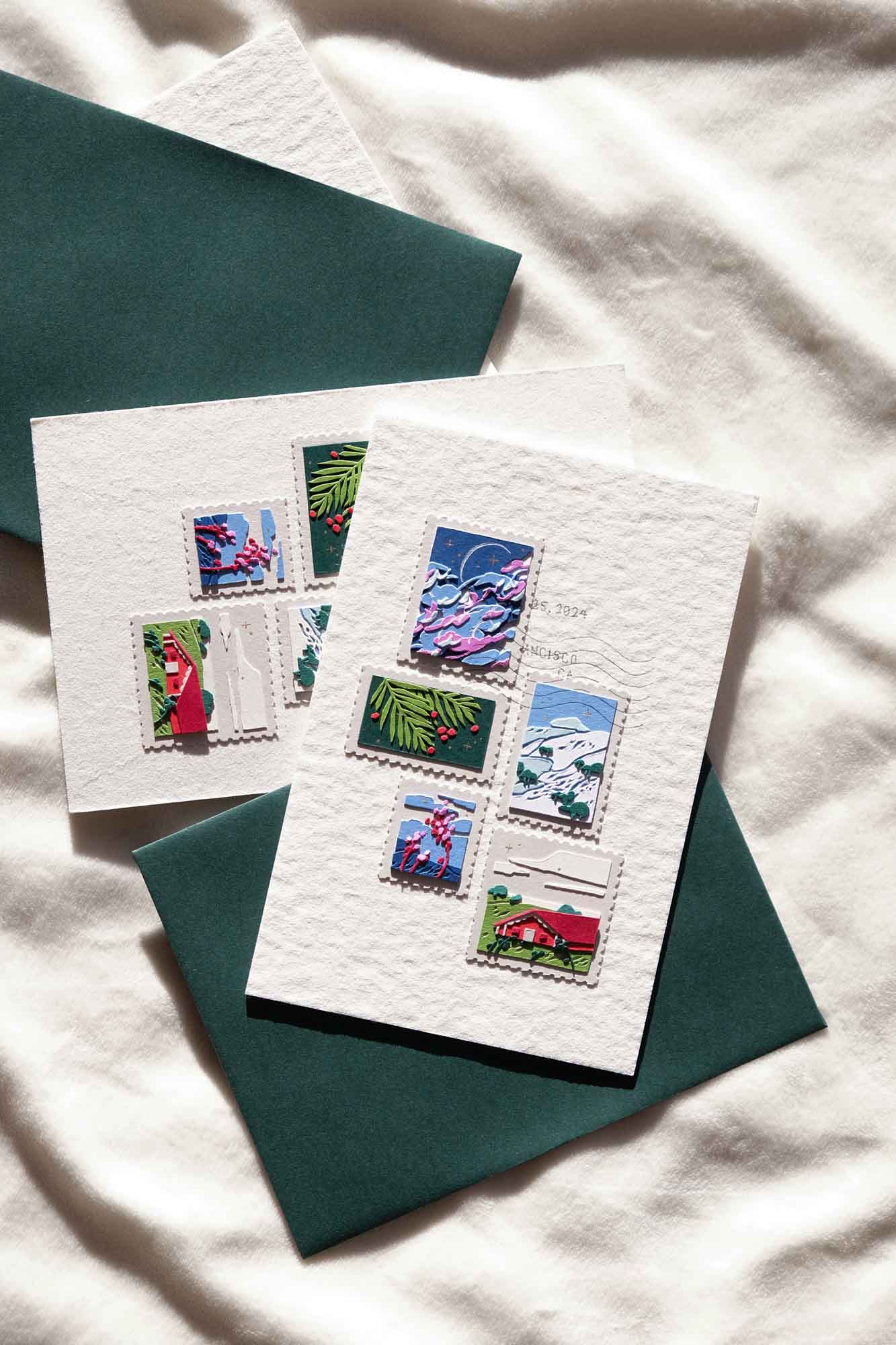 Two holiday cards, each decorated with paper stamps depicting scenes of San Francisco, sit on a snow-like surface next to deep green envelopes.