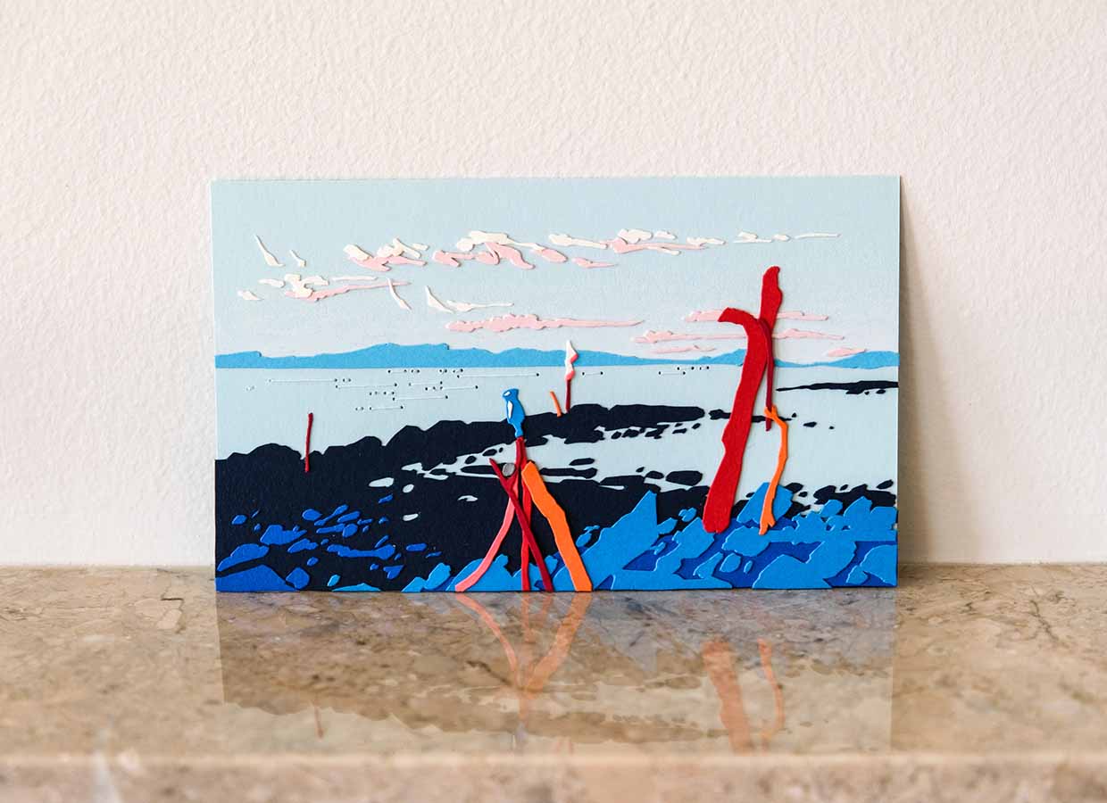 A blue paper scene of rocks, water, and distant hills is punctuated by red paper knobs of driftwood and thin paper clouds. The artwork balances on a polished stone mantle that casts a textured reflection.