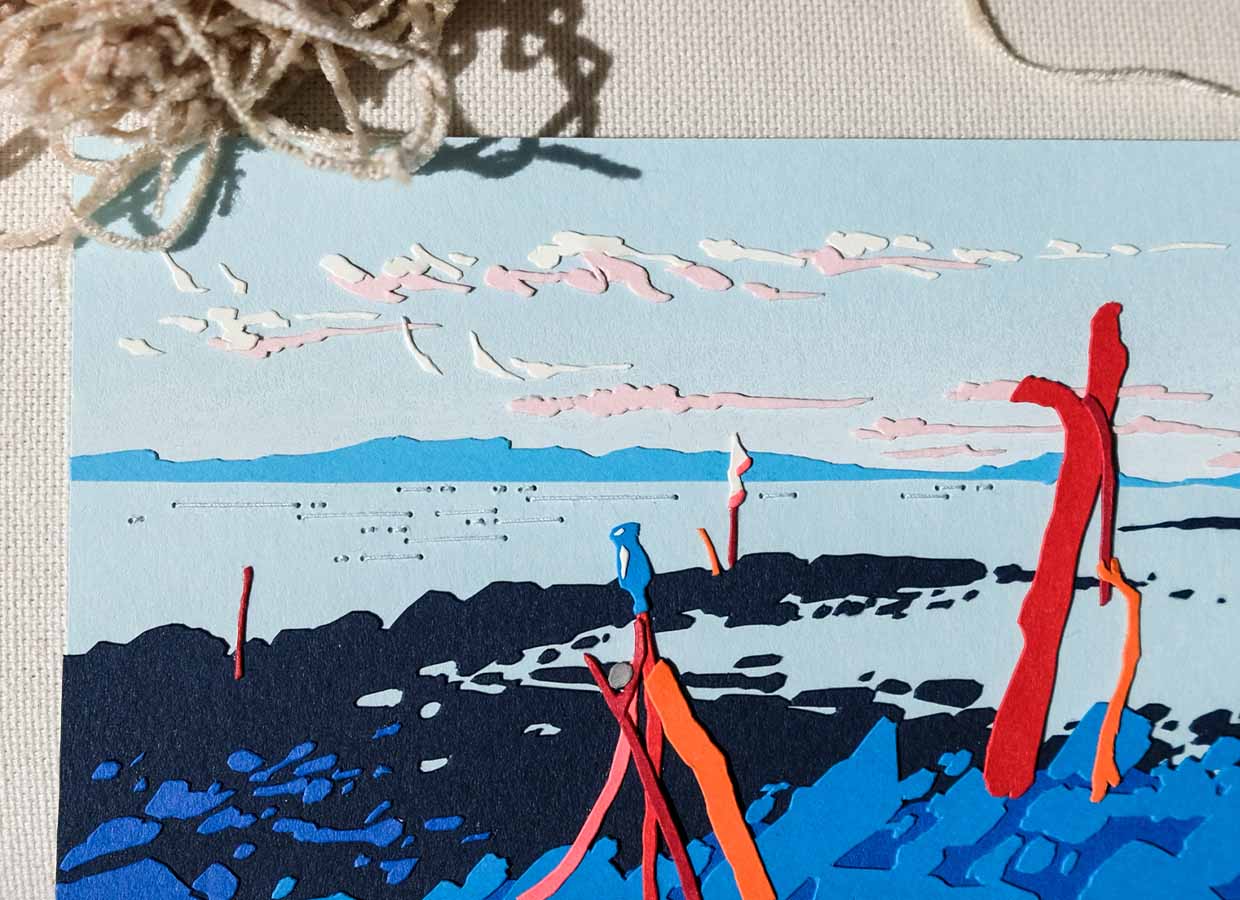 A blue paper scene of rocks, water, and distant hills is punctuated by red paper knobs of driftwood and thin paper clouds.