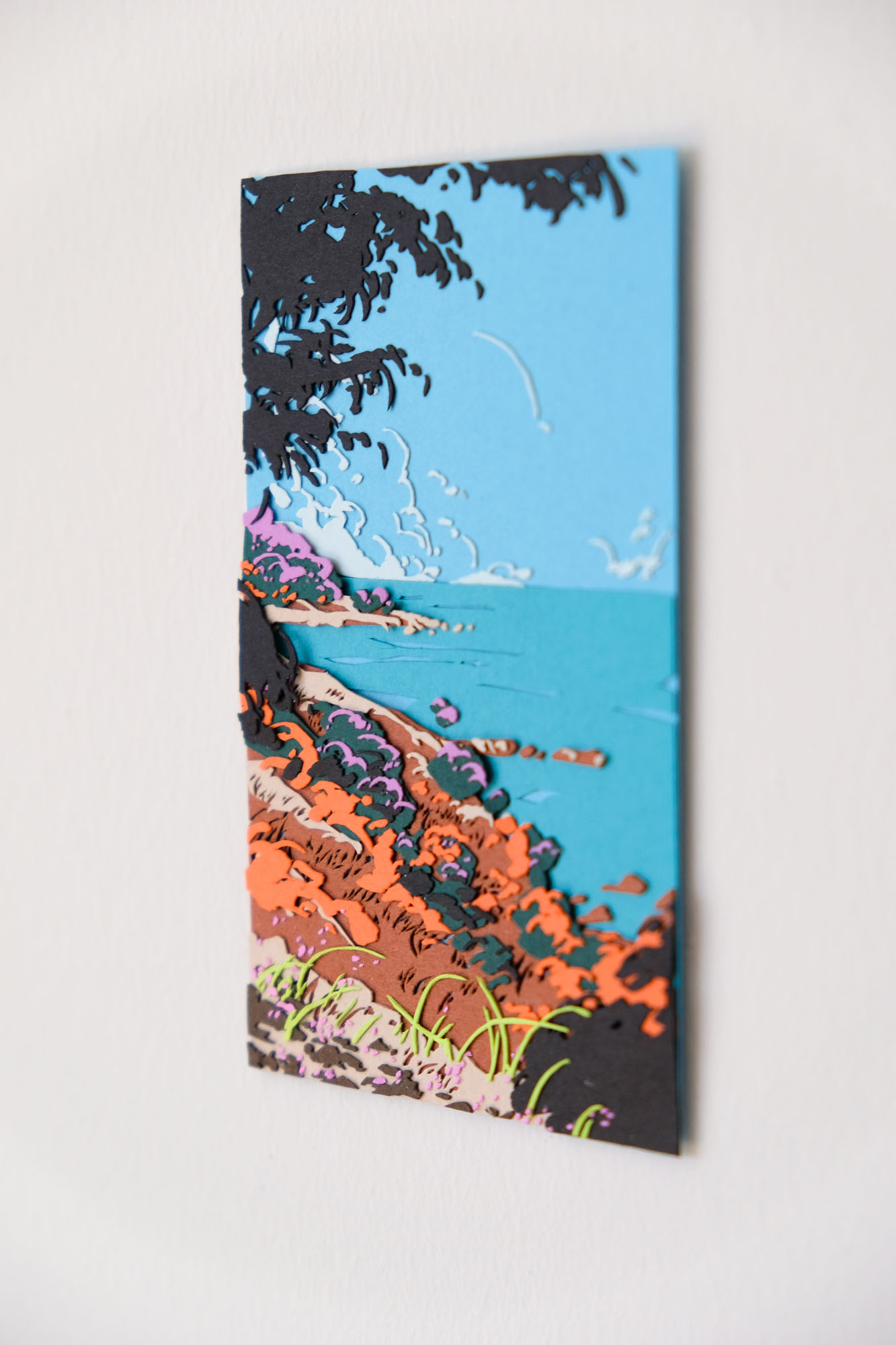 A blue ocean and a light blue sky are covered by brown paper cliffs and accents of deep green, orange, lavender purple, beige, and light green. Many of the accents evoke the forms of shrubs, foliage, and grasses.