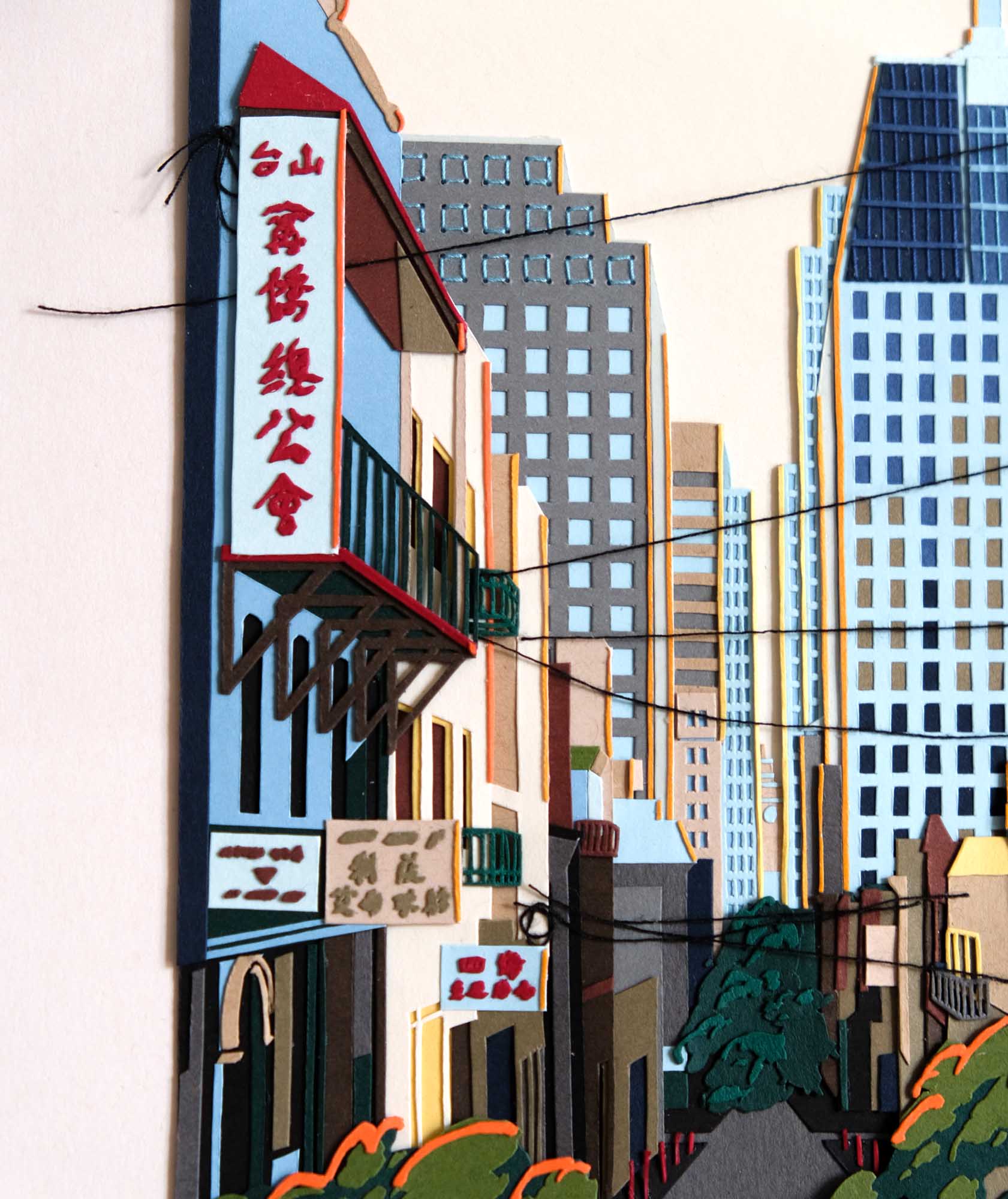 A street at the border of Chinatown and the Financial District is recreated in layers of colored papers and thread. The artwork is framed in light wood, and it sits on a hardwood floor against a vintage stepped shelf.