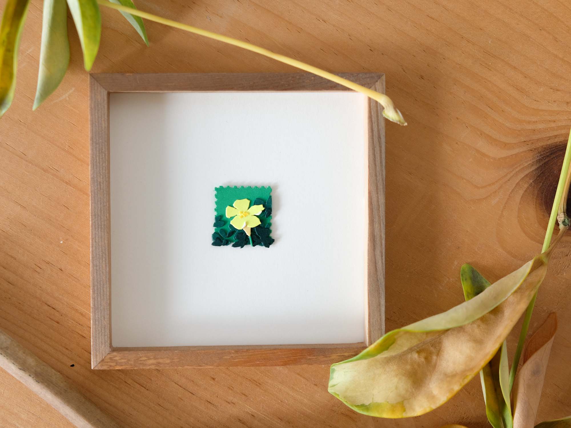 The closed yellow blooms of paper oxalis hang from the ends of graceful stems amidst a cluster of green paper leaves. The scene protrudes from a light yellow rectangle and overlaps onto a white background bordered by a dark, thin wood frame.