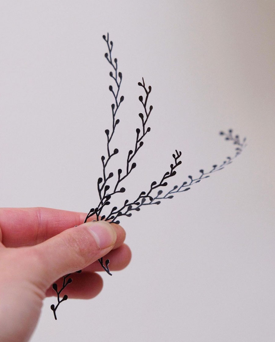 The artist's hand holds a bouquet of cut out kelp against a white wall.