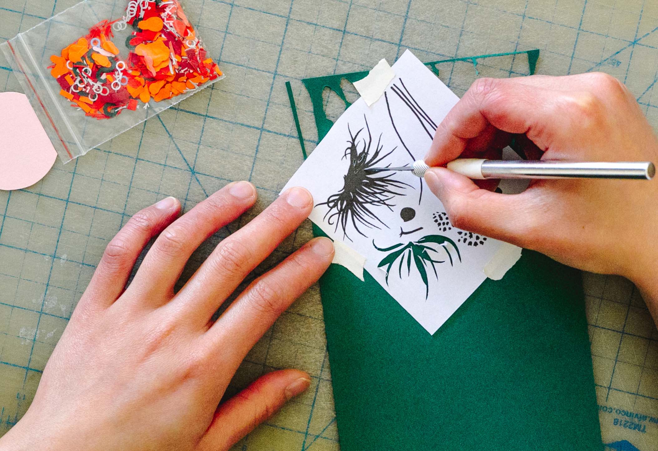 The artist's hands cut a template of leaves and flowers on a green sheet of paper using a craft knife. A small set of cut out elements sits nearby.