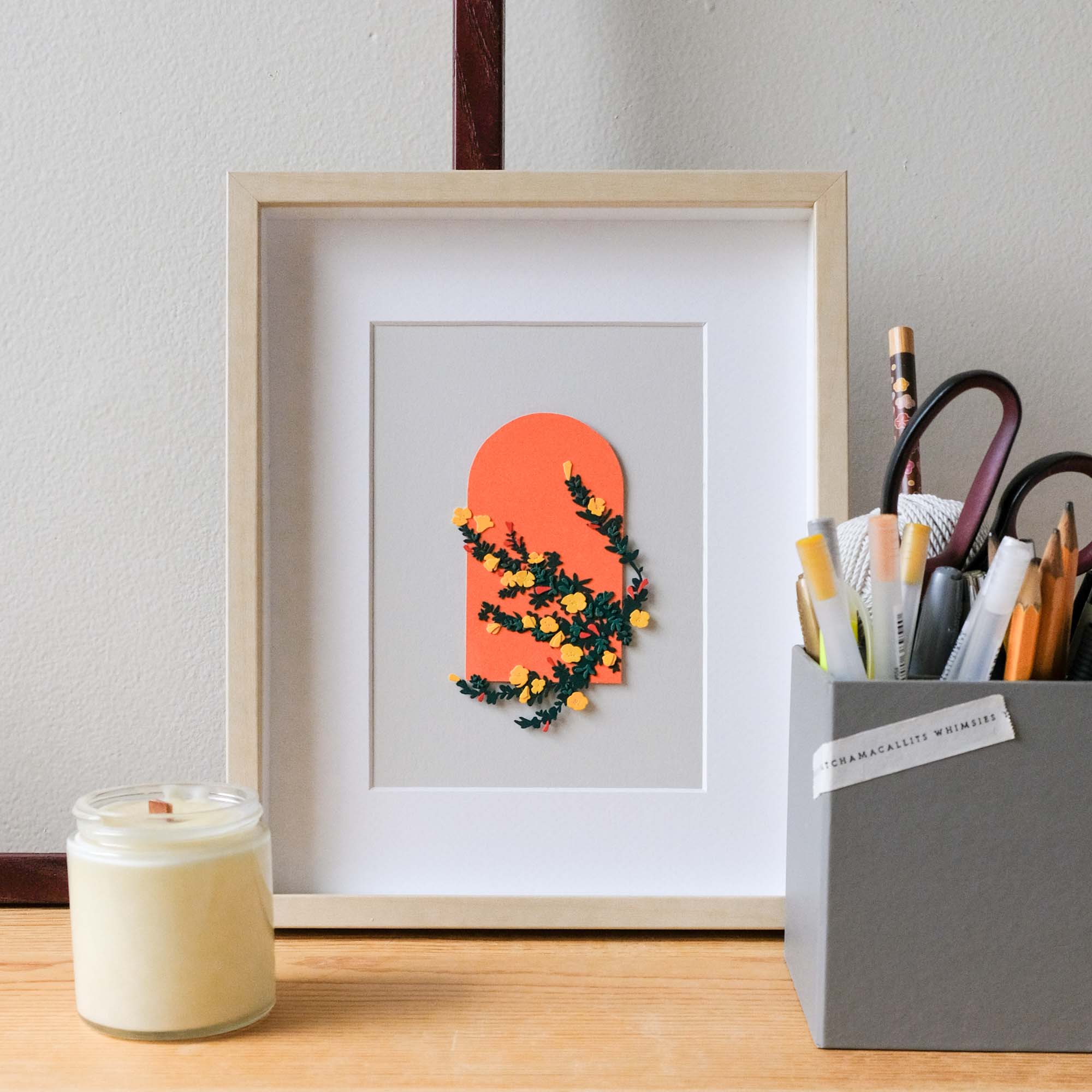 A framed copy of the paper beach primrose sits on a desk with a candle.