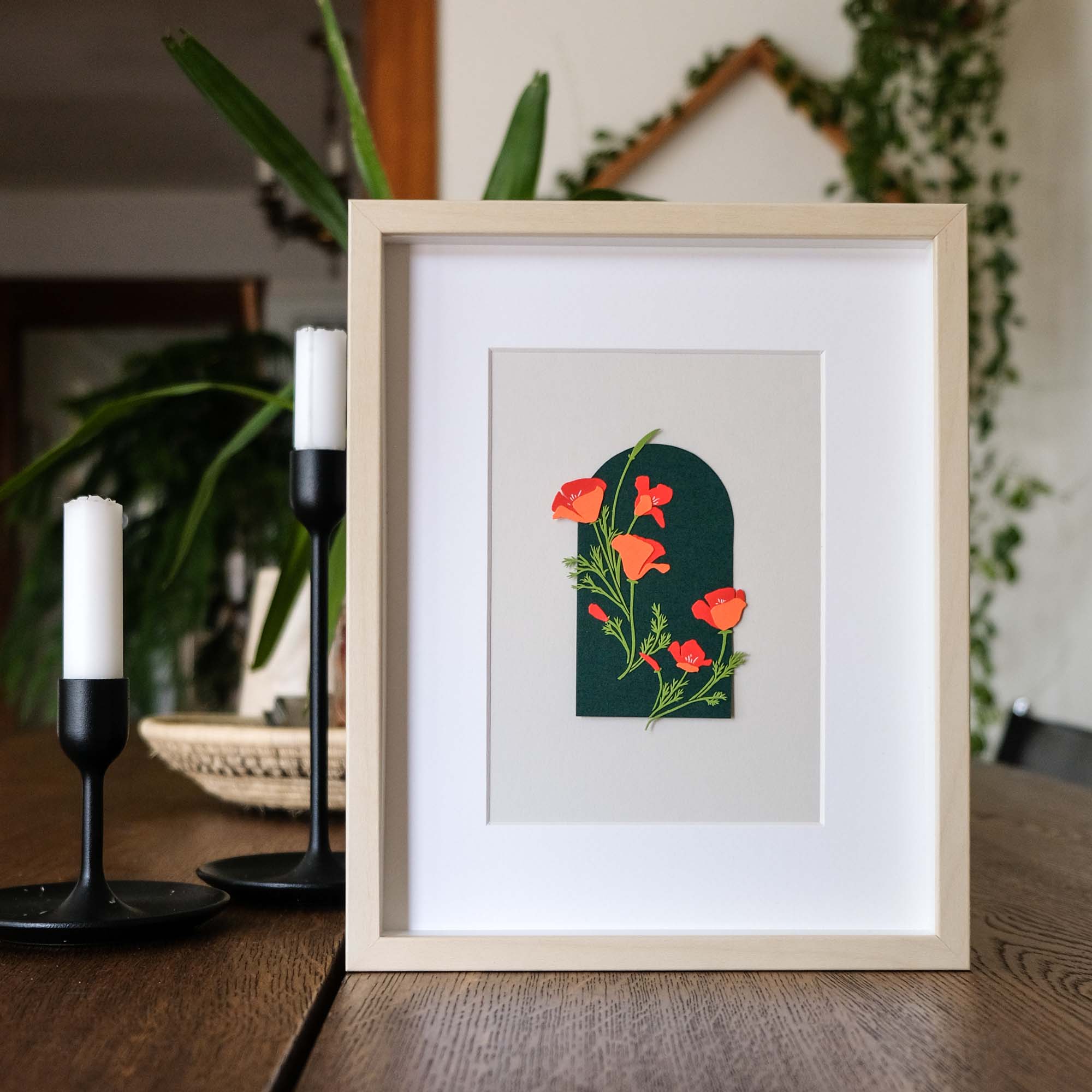 Paper California poppy flowers sit on an arched dark green background. The artwork is framed on a wood table next to candles and houseplants.