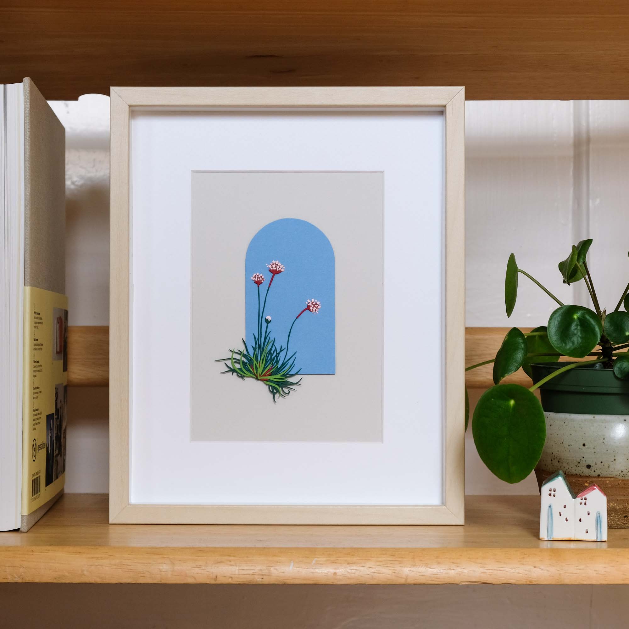 Paper sea thrift flowers sit on an arched blue background. The artwork is framed on a shelf next to a small houseplant.