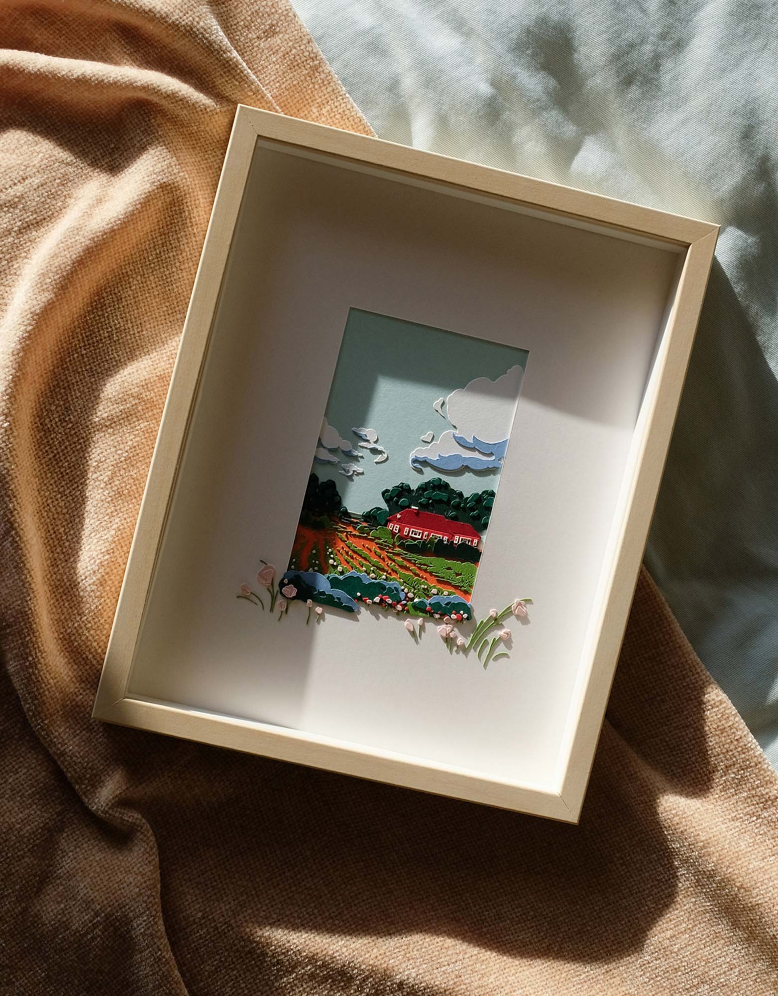 The framed artwork lies on a backrop of sky blue and neutral fabrics under strong sunlight.