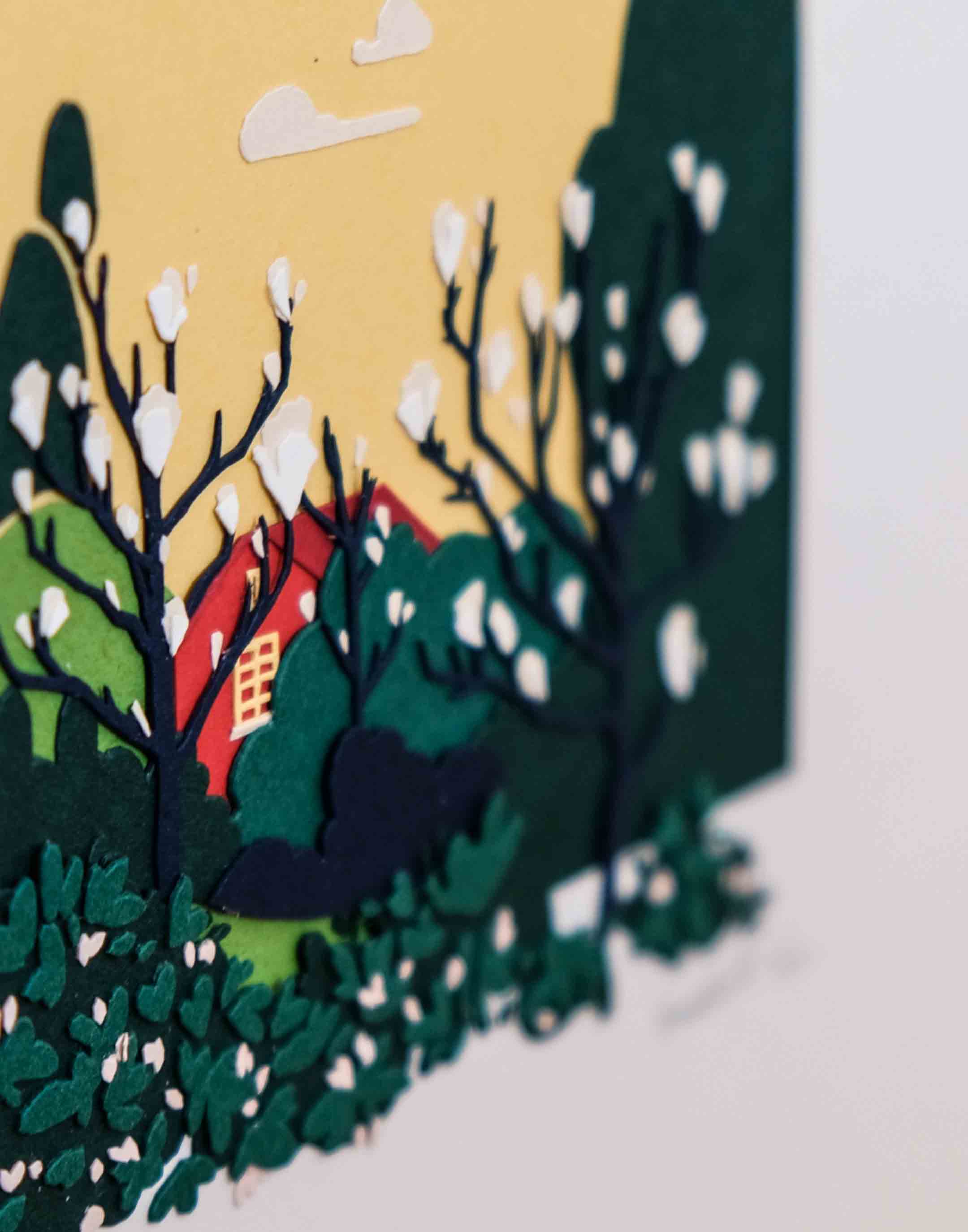 A close-up of the artwork shows the layers of paper that make up the house, magnolia blossoms, and trees.