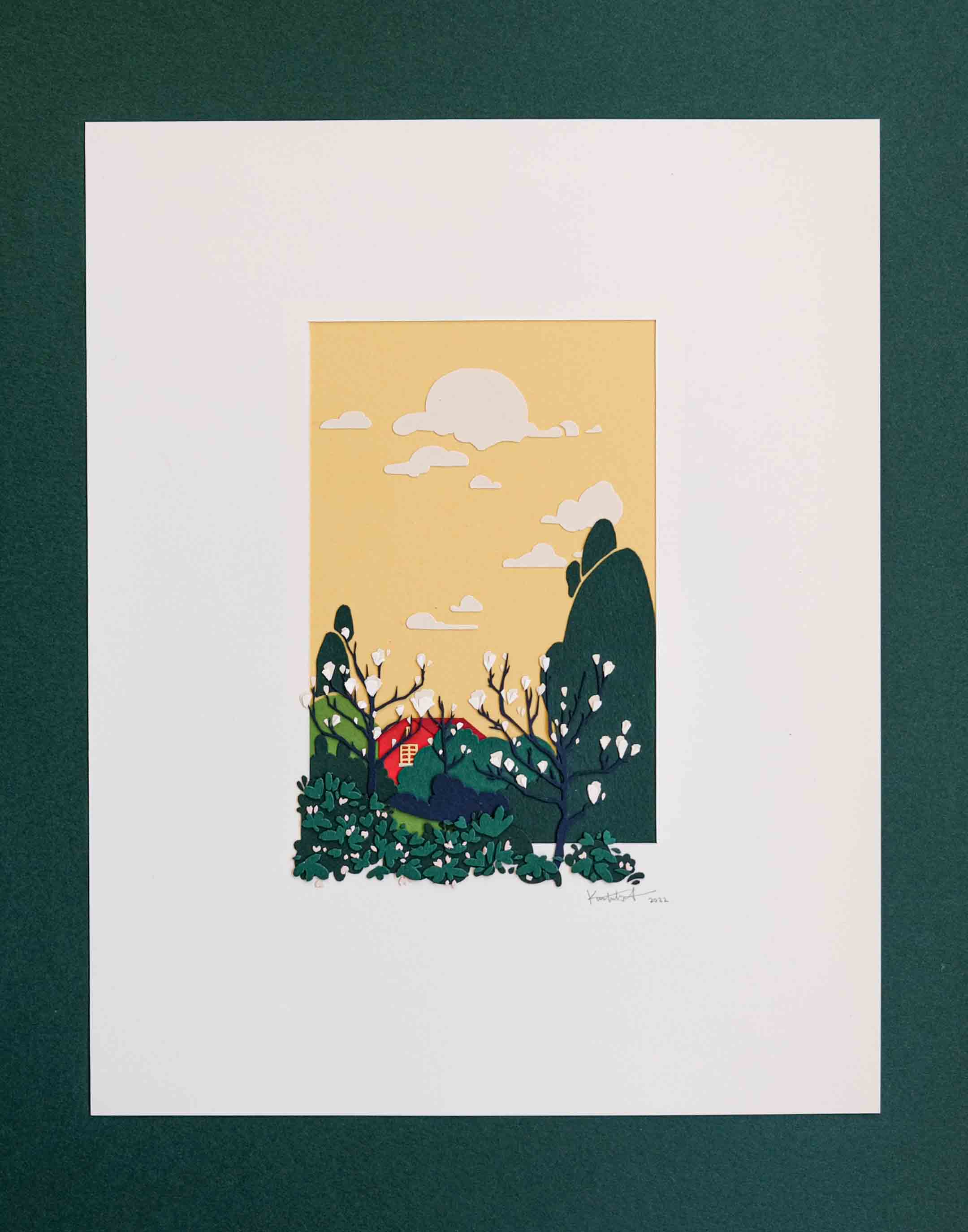Layers of cut paper depict a red house against a warm yellow sky, behind a grove of magnolia blossoms and other trees. It sits in a white mat, with some of the trees and plants spilling out of the mat opening. A dark green background is visible behind the artwork.