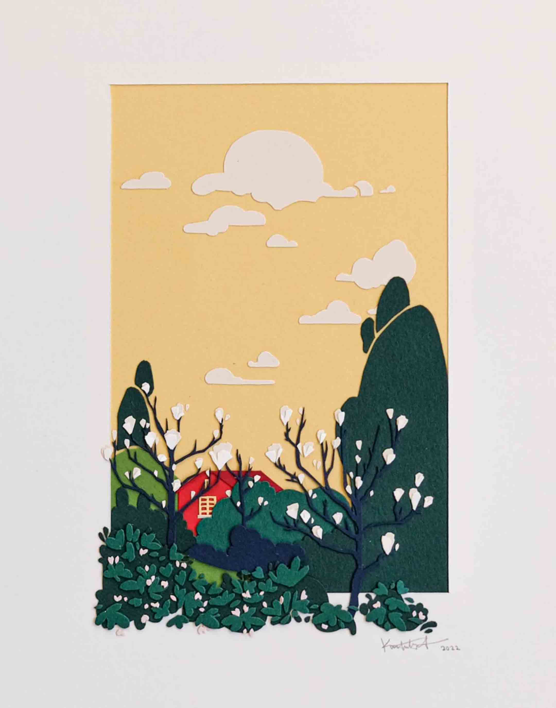 Layers of cut paper depict a red house against a warm yellow sky behind a grove of magnolia blossoms and other trees. It sits in a white mat, with some of the trees and plants spilling out of the mat opening.