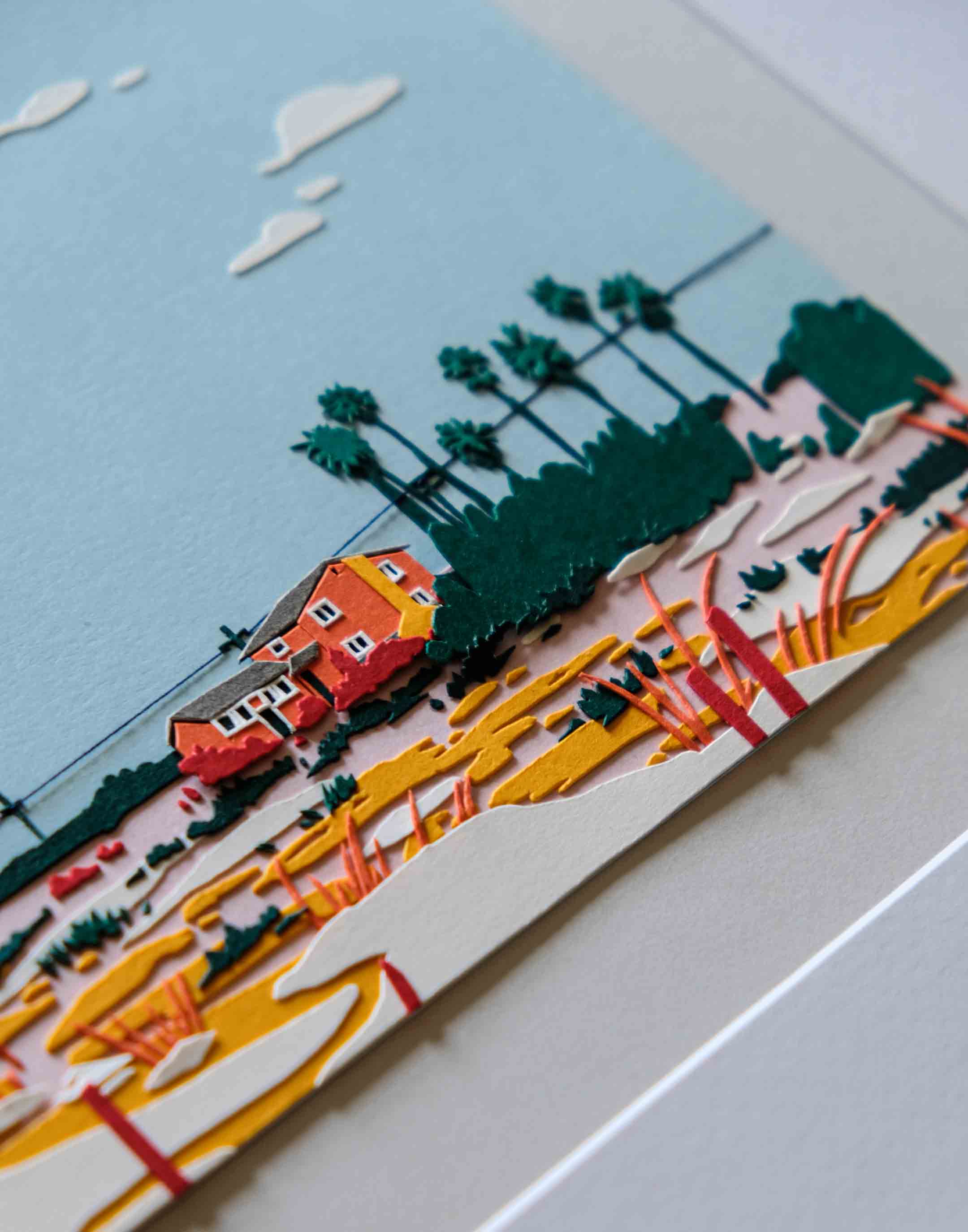 An angled close-up shows the layers of paper that form the bright orange two-story house, warm-colored fields, dark green palm trees, and white clouds.
