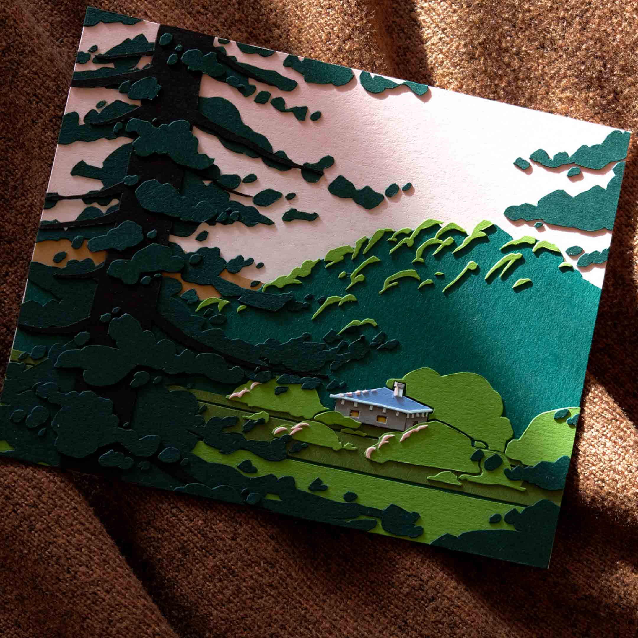 A little gray paper house sits in a green paper field, peeking between paper trees during sunset. The artwork sits partially in a shaft of sunlight on a warm cloth background.