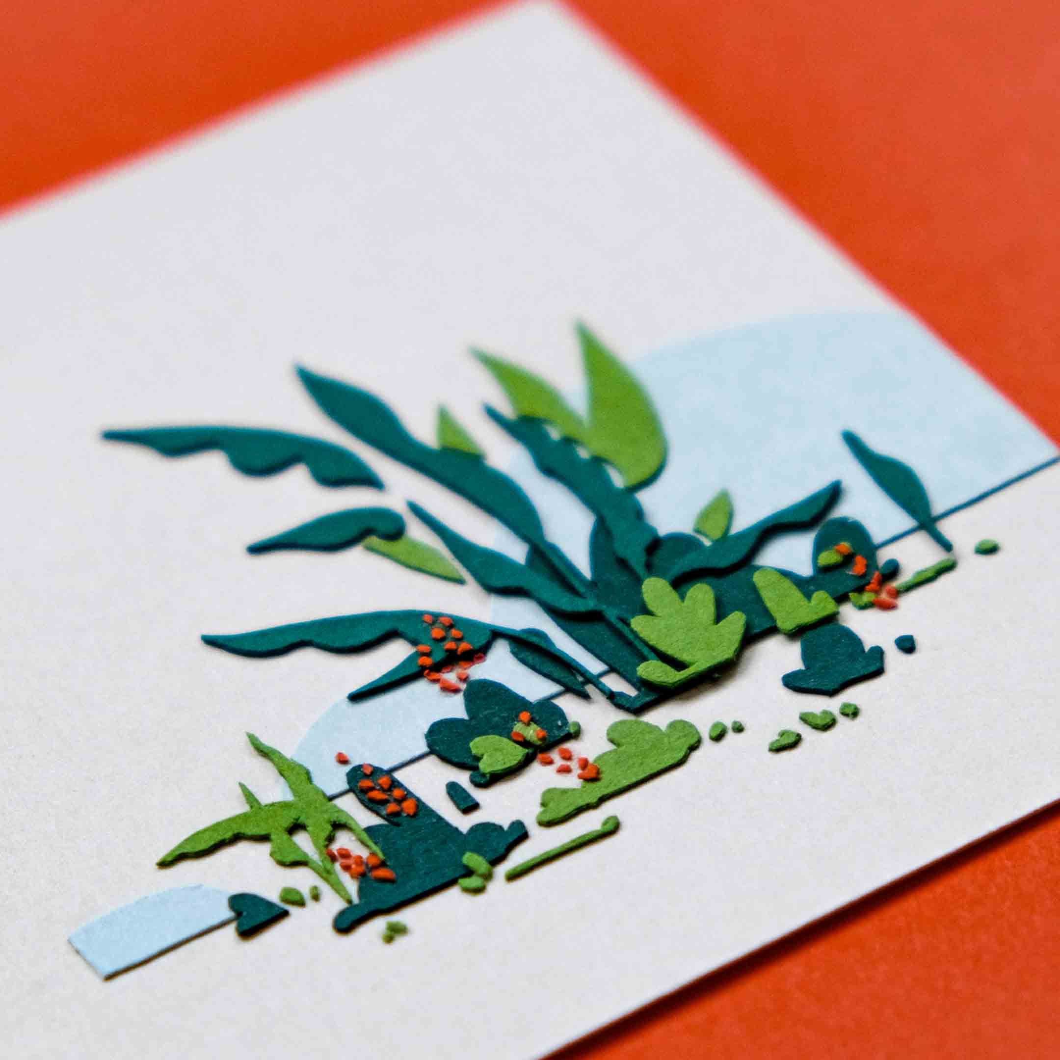 An angled view of a small square artwork shows layers of green paper tropical plants in front of blue paper hills. The artwork is on an orange backdrop.